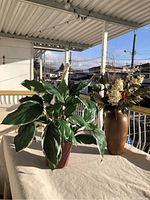 Two items on a covered outdoor table: a live leafy plant in a red pot and a tall brown vase with an artificial floral arrangement.