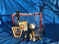 Assorted sports equipment displayed on blue tarp including mini goal/net, rackets, soccer ball, shuttlecocks, stopwatch, hand grip strengthener, and air pump.