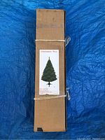 Boxed artificial Christmas tree tied with strings, with a label showing tree image and product details.