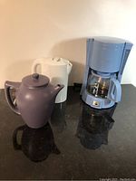 Photo showing all three items: purple ceramic teapot, white electric kettle, and light blue coffee maker with glass carafe on black countertop.