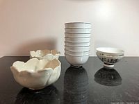 Display of all bowls: seven stacked white bowls with black rim line, two larger scalloped white bowls, and two smaller character-decorated bowls.