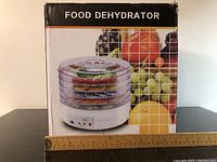Photo showing the front of the food dehydrator box with ruler placed beneath for scale, featuring the product image and label 'FOOD DEHYDRATOR.'