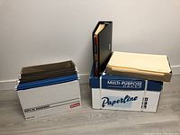Hanging file folders in blue, green, and manila stacked in a box and manila file folders stacked beside a large black binder with tabs visible.