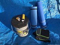 Photo showing Reliance portable toilet with attached handle, yellow and black flashlight, silver and black flashlight, and two rolled blue camping mats on blue tarpaulin.