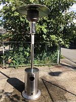 Full view of Nexgrill stainless steel patio heater standing outdoors on a concrete surface, showing base, pole, burner area and heat reflector.