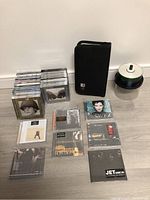 Photo showing a collection of various music CDs arranged on the floor, including jewel-cased albums and paper sleeve CDs, a large spindle stack of blank recordable CDs, and a black zippered CD storage case.