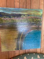 Image showing an unframed, unsigned abstract landscape oil painting on stretched canvas. The painting features layered colors including blue, green, yellow, purple, and brown, creating a textured landscape scene with a large blue area resembling water. The canvas appears lightly used with some gloss from the paint.