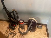Photo showing Bissell upright vacuum and Progressive canister vacuum on floor against the wall, with cords and hoses visible.
