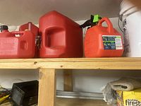 Photo showing four lined-up red plastic gas containers on wooden shelf, different sizes and shapes, clean and empty.