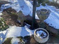 Photo showing two large planters in a snowy garden area, one urn-shaped with vertical swirl design and one round planter with decorative geometric patterns.
