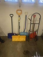 Five items comprising four snow shovels of different sizes and a metal dolly. The snow shovels have plastic blades in black, yellow, blue, and orange, all with D-shaped handles. The dolly has a red frame with two black wheels and a foot plate.
