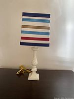 White table lamp with twisted column base and colorful striped shade photographed on a flat surface against a plain wall.
