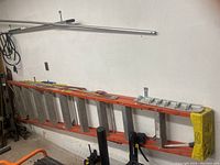 Full view of tall aluminum ladder with orange frame placed on ground leaning against a wall.