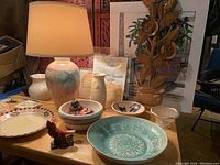 Photo showing a ceramic table lamp, various ceramic bowls and plates, small figurines, and framed watercolors arranged on a wooden surface.