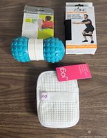 Photo showing three items: blue Gaiam dual zone back roller with textured surface, Gozone fabric squat band in box, and Jogi white exfoliating bath pad with strap and tag.