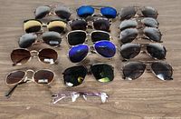 Photo showing all 15 sunglasses and one pair of eyeglasses laid out on a wooden surface, various frame styles and lens colors visible.