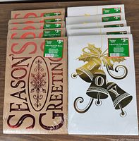 Photo showing two distinct styles of holiday gift boxes: one design features "Season's Greetings" text in red on a beige background, the other shows gold bells with black and gold accents and the word "Joy" on a white background. Both sets have green price stickers labeled "Holiday Style Value Pack Gift Boxes".