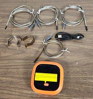 Top-down photo of the thermometer unit with four probes, two metal clip holders, and USB charging cable arranged on wood surface.