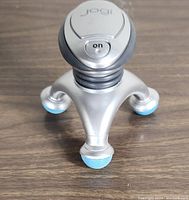 Close-up of the silver Jogi handheld massager with three legs and blue rounded tips, showing the on/off button on top with Jogi branding.
