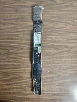 Single black metal mulching blade packaged in clear plastic on wood surface, label visible