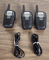 Photo of three black Uniden SX307-3C two way radios with antennas and display screens, placed side by side on a wooden surface with two black charging cables in front.