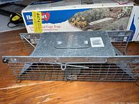 Live cage trap for small squirrels placed on a wooden surface with box in background, showing the metal cage and handle on top.