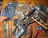 Photo showing assorted hand tools including hammer, door lock set, various metal garden tools and trowels on wooden surface