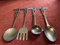 Four brass cooking utensils hung on a wall mount rack, displayed on red carpet background. Utensils include skimmer spoon, fork, ladle, and solid spoon, all about 15 inches long with decorative handles.