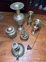 Photo of all brass items arranged on wooden table, showing embossed tall vase, smaller vases, candle holder with snuffer, lidded jar, gong, and musical swan figurines