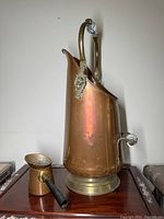 Copper coal scuttle with brass base ring, white ceramic handle grips, and small accompanying copper cooking pot with black handle shown from front-angle view.