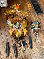 Two cuckoo clocks displayed on wooden floor, showing the larger wooden clock with painted leaves and bird next to the smaller colorful clock with figurine and key.