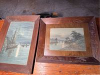 Two large paintings framed in wood, showing one scene of sailboats on water and the other a river with bridge and cabin, with dusty glass and visible wood texture on frames
