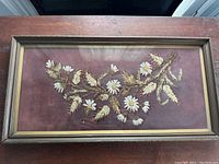 Framed rectangular embroidery artwork showing a floral design with white daisies and brownish-golden leaves on a burgundy fabric background.