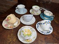 Seven assorted English cup and saucer sets arranged on a surface, showing different floral and decorative patterns.