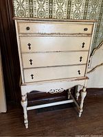 Front view of the antique French Provincial style highboy, showing peeling cream paint and four drawers with metal knobs and keyhole escutcheons.