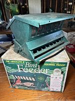 Green metal squirrel proof bird feeder sitting on its original box exhibiting adjustable feeding slots and squirrel proof design