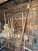 A collection of long-handled tools including pitchforks, garden rakes, shovels, and rusted ice tongs hanging on a rustic wooden wall.