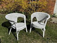 Two white wicker chairs positioned on outdoor grass with a brick wall and yellow foliage in background. View is slightly angled showing front and side of chairs.