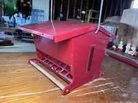 Red metal bird feeder on a wooden surface showing front and side with wooden perch
