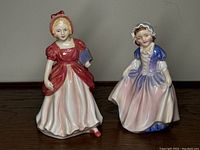 Two small porcelain figurines, one in a blue and white dress and bonnet, the other in a red and white dress holding a book.