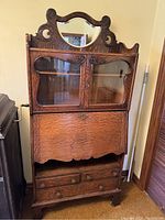 Front view of the tall drop front desk bookcase showing the oak wood grain, glass-front cabinet doors with hardware, and decorative crest with bevelled mirror.