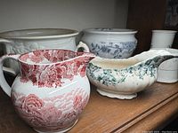 Six items including two chamberpots, a red transferware pitcher, a blue-green floral cream boat, a small cup and a tall cup with blue-green decoration on a wooden shelf.