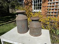 Two vintage metal milk cans on a white table outside next to brick wall and window. One shorter, embossed with Swift, the other taller with faint stamp. Both have lids and side handles.