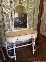 Front view of antique French provincial vanity showing curved top and drawer, ornate mirror, turned legs, and flaking paint.