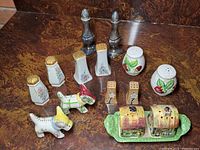 Vintage salt and pepper shakers in various styles displayed on a dark wood surface: includes floral ceramic, metallic pair, strawberry ceramic shakers, two dog figurine shakers, and a ceramic cottage house pair with tray.