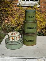 Full set of four graduated green tins stacked labeled TEA, SUGAR, FLOUR and two additional decorative tins stacked next to them on white table outdoors.