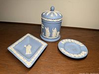 Three pieces of Wedgwood jasperware including covered dish, square tray, and round tray, showing details of white relief classical figures on blue background.