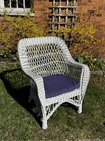Front side view of white wicker armchair with removable blue seat cushion placed outdoors on grass with brick wall background.