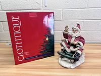 Clothtique Santa figurine seated on sled next to original red box with branding and Christmas tree illustration.