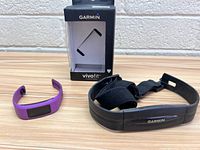 Garmin fitness tracker watch in purple band next to Garmin black heart rate monitor chest strap and boxed vivofit packaging.
